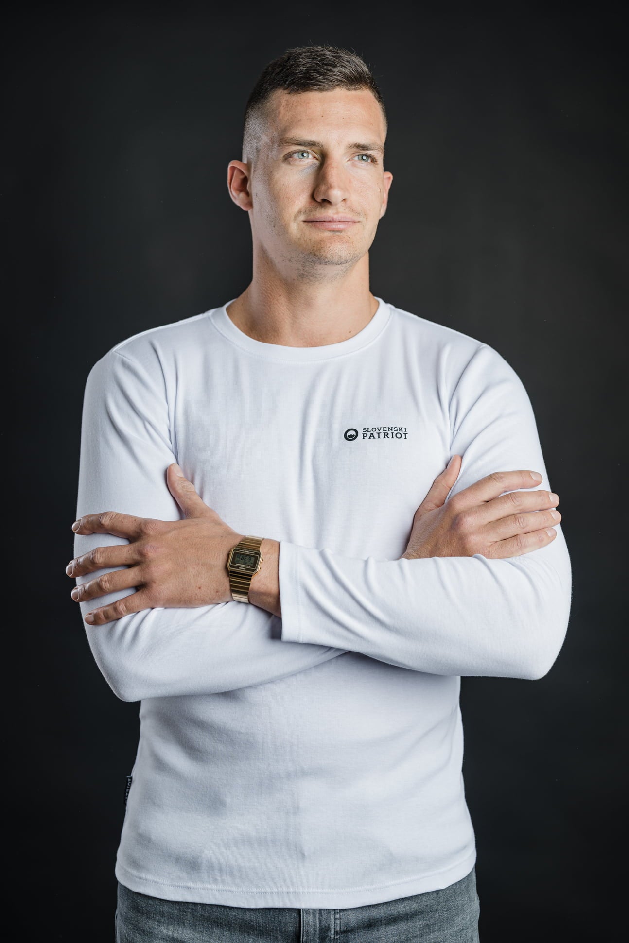 Moški s prekrižanimi rokami nosi belo 100% bombažno majico z dolgimi rokavi Tech Pro znamke Slovenski Patriot, prikazano iz bočnega kota, narejeno v Sloveniji.