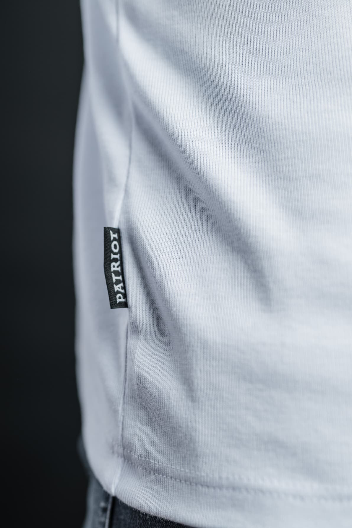 Detajl črne etikete z napisom PATRIOT, všite v stranski rob bele majice z dolgimi rokavi TECH PRO, narejene v Sloveniji.