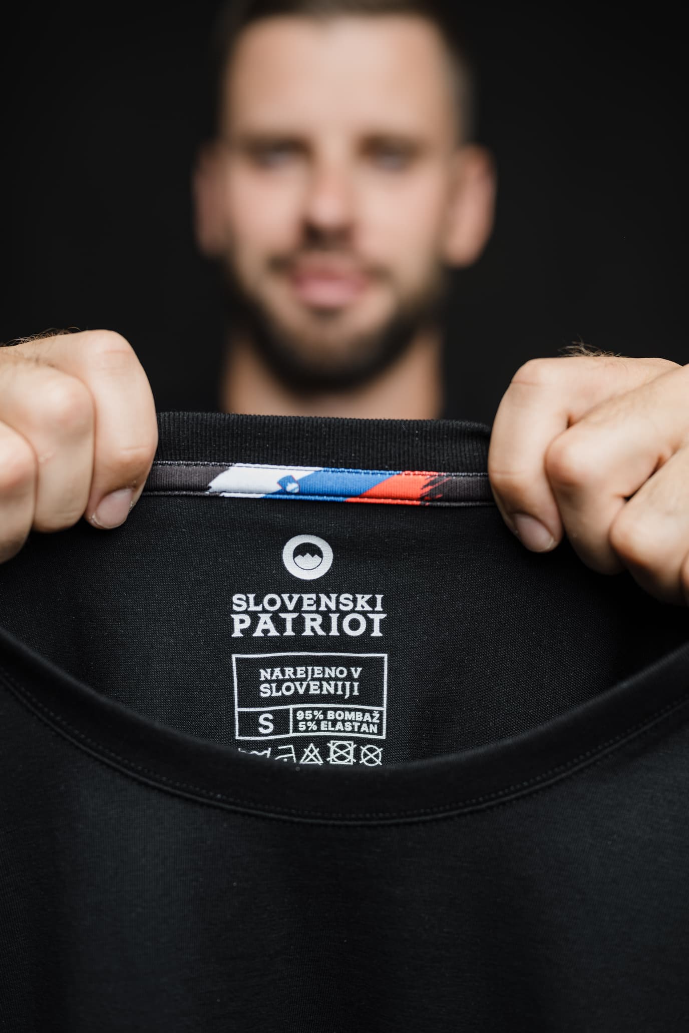 Detajl notranje etikete črne majice FIT znamke Slovenski Patriot, z napisom "Narejeno v Sloveniji", oznako sestave in slovenske zastave.