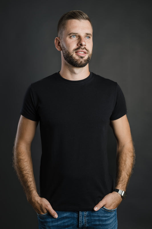 Moški v črni majici FIT znamke Slovenski Patriot stoji sproščeno z rokami v žepih in gleda vstran; majica poudari oprijet kroj in minimalističen dizajn narejen v Sloveniji.