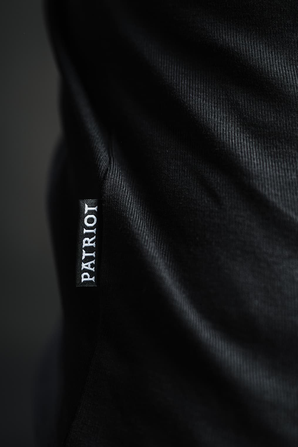 Detajl črne etikete z napisom PATRIOT, všite v stranski rob črne majice z dolgimi rokavi TECH PRO, narejene v Sloveniji.