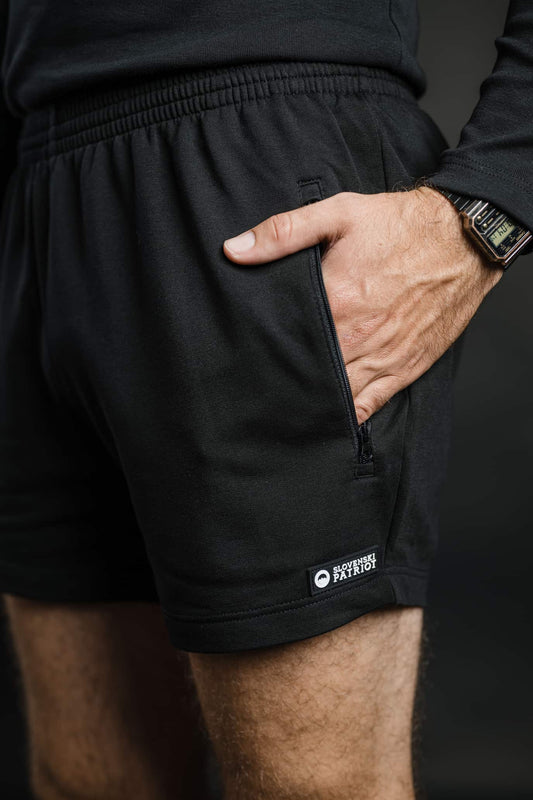Moški ima roko v žepu črnih kratkih hlač MOVE znamke Slovenski Patriot, ki imajo elastičen pas in zadrgo, narejene iz bombaža v Sloveniji.