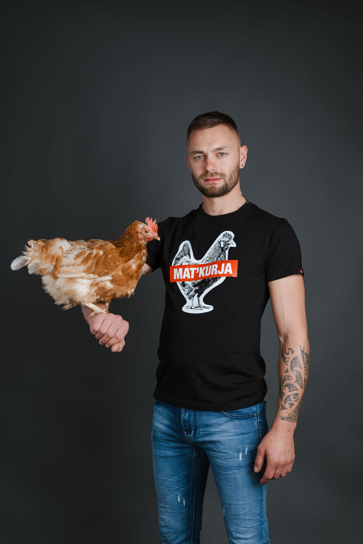 Moški v jeans hlačah drži kokoš in nosi črno bombažno majico Matkurja znamke Slovenski Patriot, narejeno v Sloveniji.