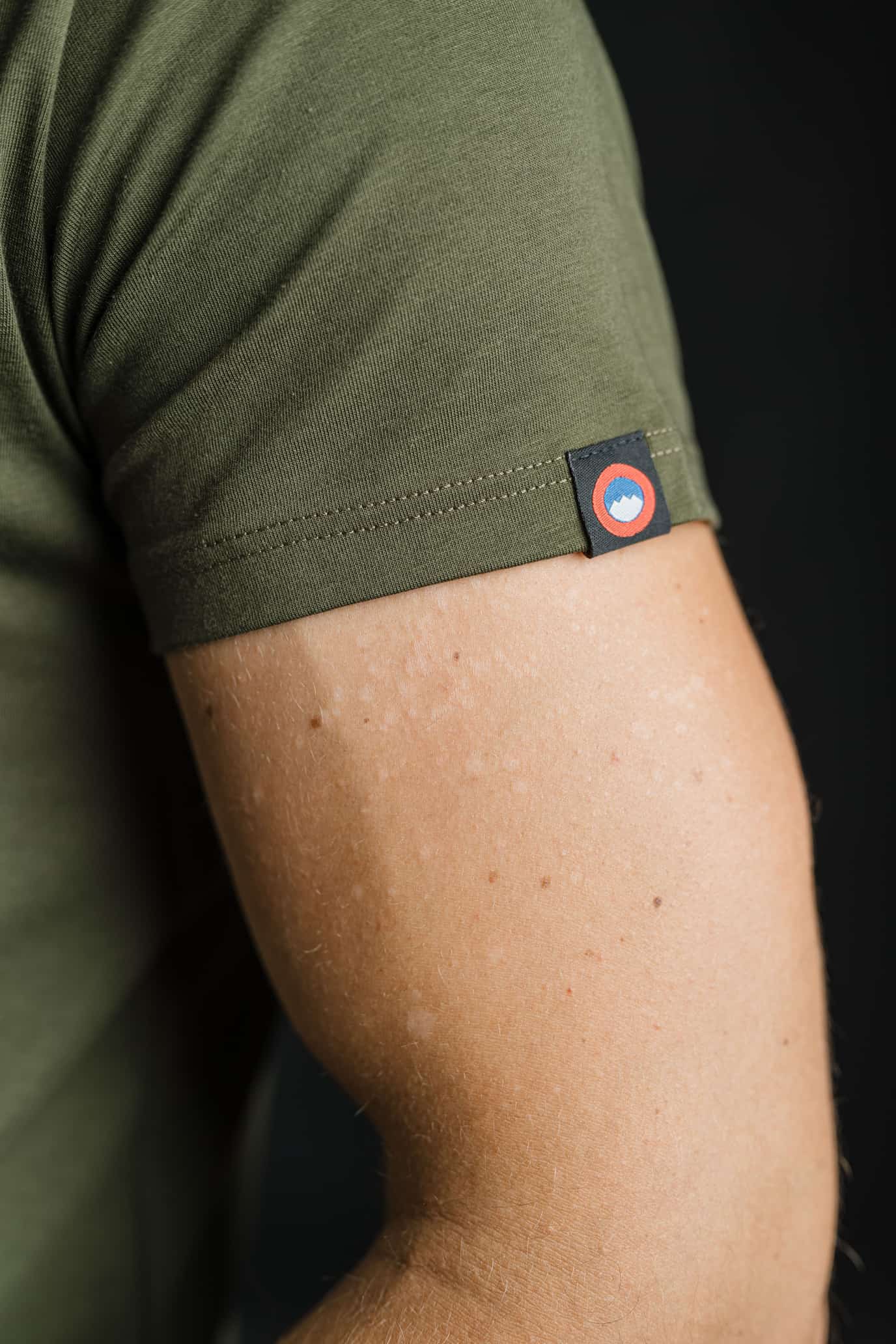 Bližnji posnetek rokava olivne majice FIT znamke Slovenski Patriot z všitim logotipom v rdeče-modri barvi, ki poudarja pozornost do detajlov in slovensko identiteto.