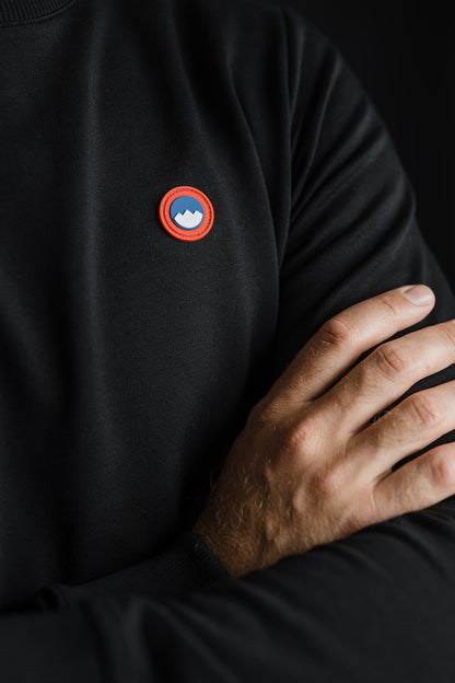 Detajl črnega puloverja Patriot Crew znamke Slovenski Patriot, z vidnim rdeče-modrim logotipom na prsih in moško roko prekrižano čez prsi, narejen v Sloveniji iz kakovostnega bombaža.