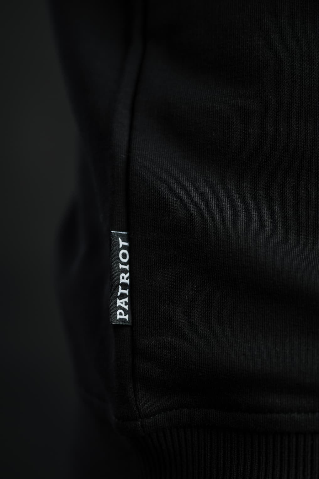 Detajl črne etikete z napisom PATRIOT, všite v stranski rob črnega puloverja Patriot Crew znamke Slovenski Patriot, narejenega v Sloveniji.