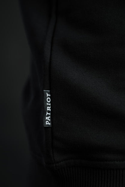 Detajl črne etikete z napisom PATRIOT, všite v stranski rob črnega puloverja Patriot Crew znamke Slovenski Patriot, narejenega v Sloveniji.