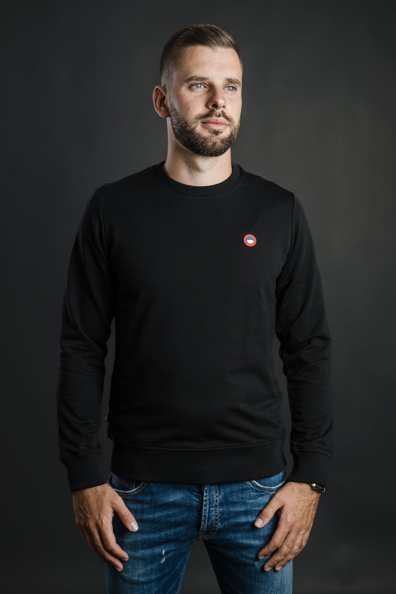 Moški nosi črn pulover Patriot Crew znamke Slovenski Patriot z rdeče-modrim logotipom, narejen iz kakovostnega bombaža v Sloveniji.