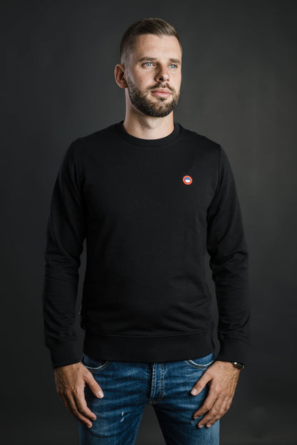 Moški nosi črn pulover Patriot Crew znamke Slovenski Patriot z rdeče-modrim logotipom, narejen iz kakovostnega bombaža v Sloveniji.