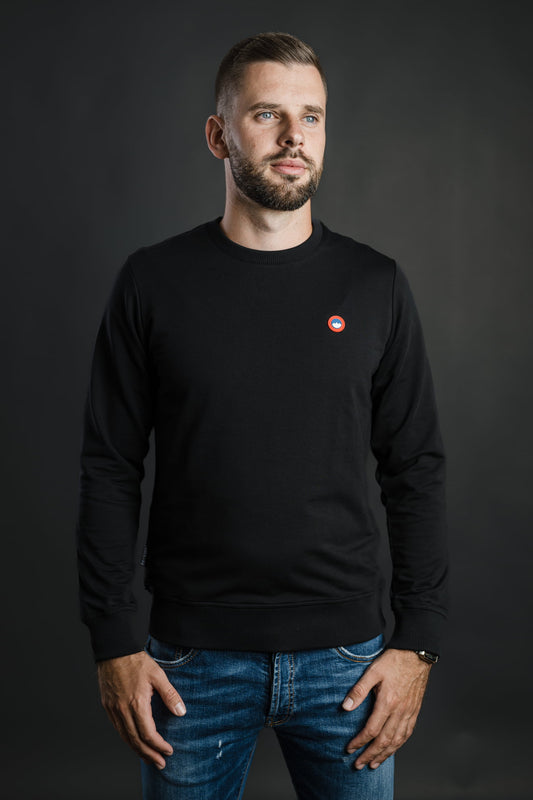 Moški nosi črn pulover Patriot Crew znamke Slovenski Patriot z rdeče-modrim logotipom, narejen iz kakovostnega bombaža v Sloveniji.