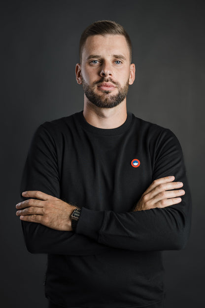 Moški nosi črn pulover Patriot Crew znamke Slovenski Patriot z rdeče-modrim logotipom na prsih, stoji z rokami prekrižanimi na prsih, narejen v Sloveniji iz kakovostnega bombaža.