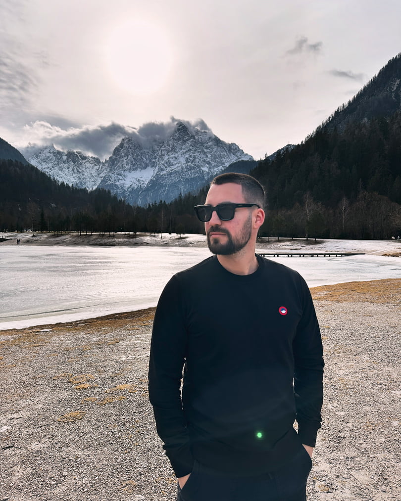 Moški nosi črn pulover Slovenski Patriot ob jezeru s pogledom na zasnežene slovenske gore. Simbol ponosa in Patriotizma.